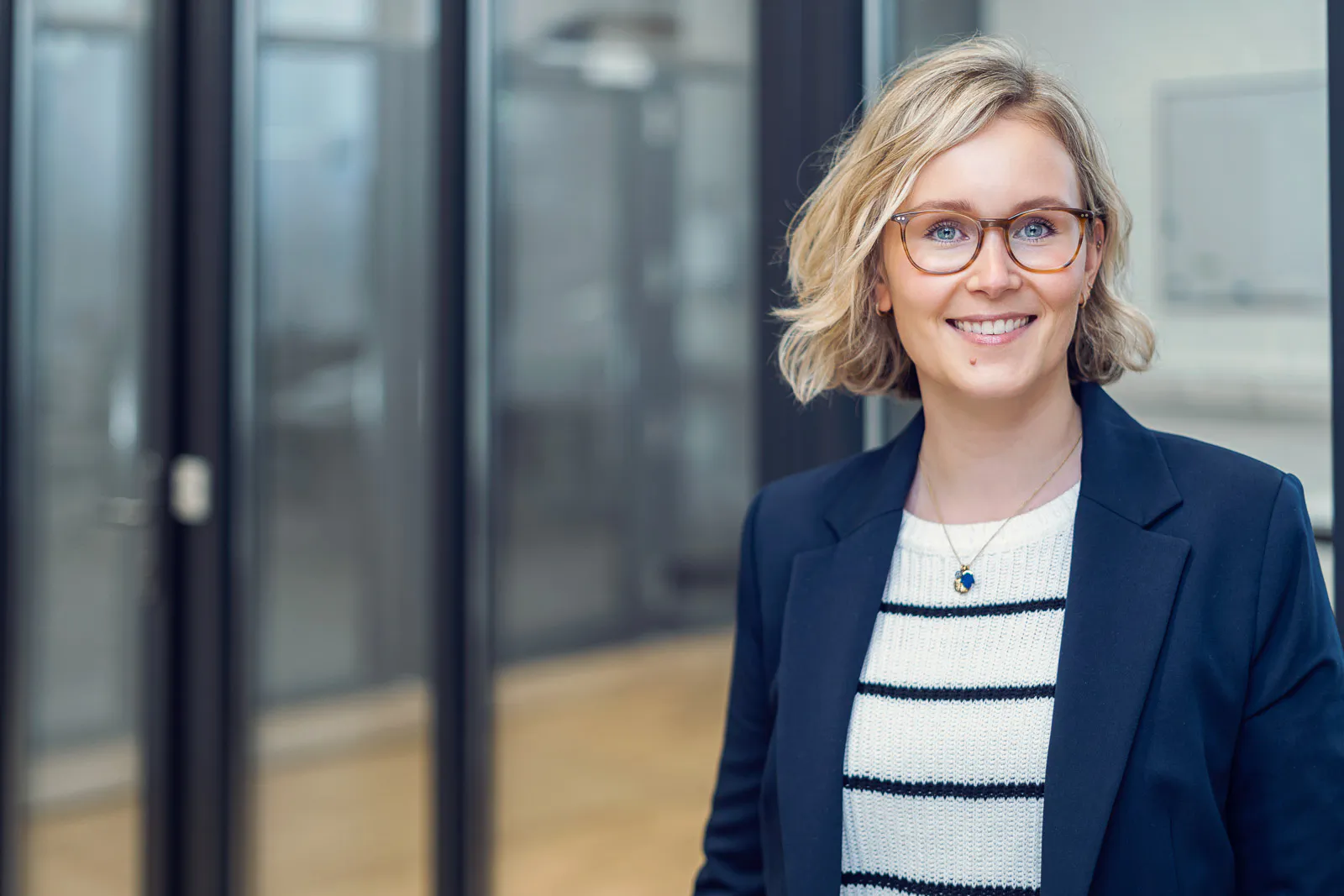 Lächelnde Frau mit Brille und dunkelblauem Blazer vor Glaswänden im Büro, in weichem Licht.