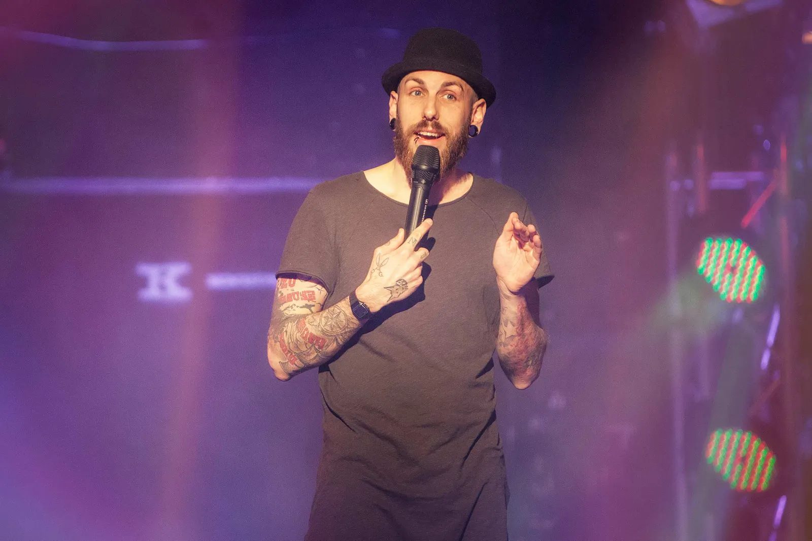Dennis Grundt mit schwarzem Hut und Mikrofon auf der Bühne im violetten Licht beim NDR Comedy Contest.