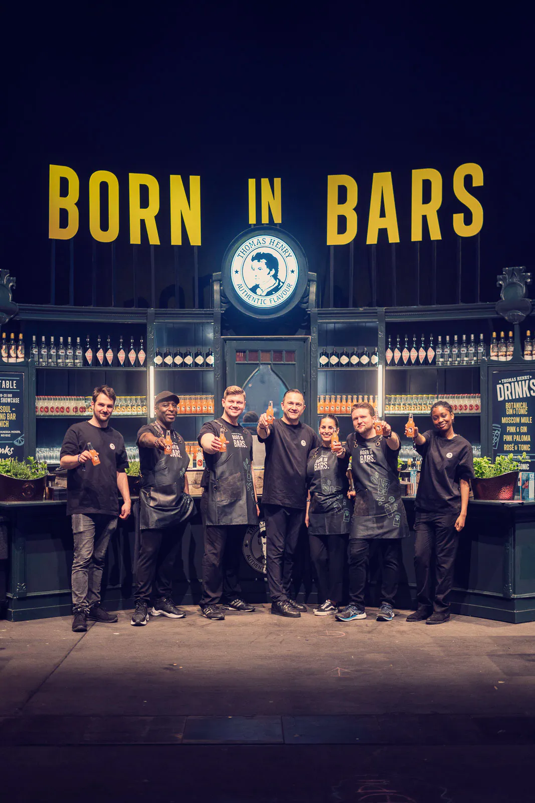 Barteam posiert vor einer Thomas-Henry-Bar mit Flaschenregalen und gelbem Schriftzug BORN IN BARS.
