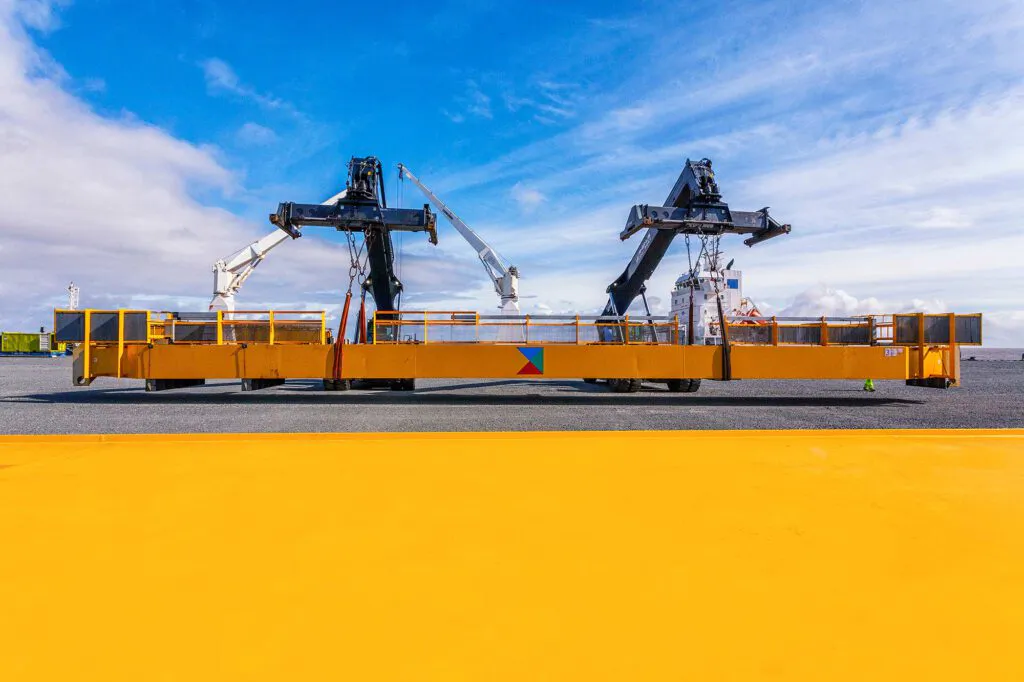 Zwei Reachstacker halten eine gelbe Lasttraverse auf einem Industriegelände unter blauem Himmel mit Schleierwolken.