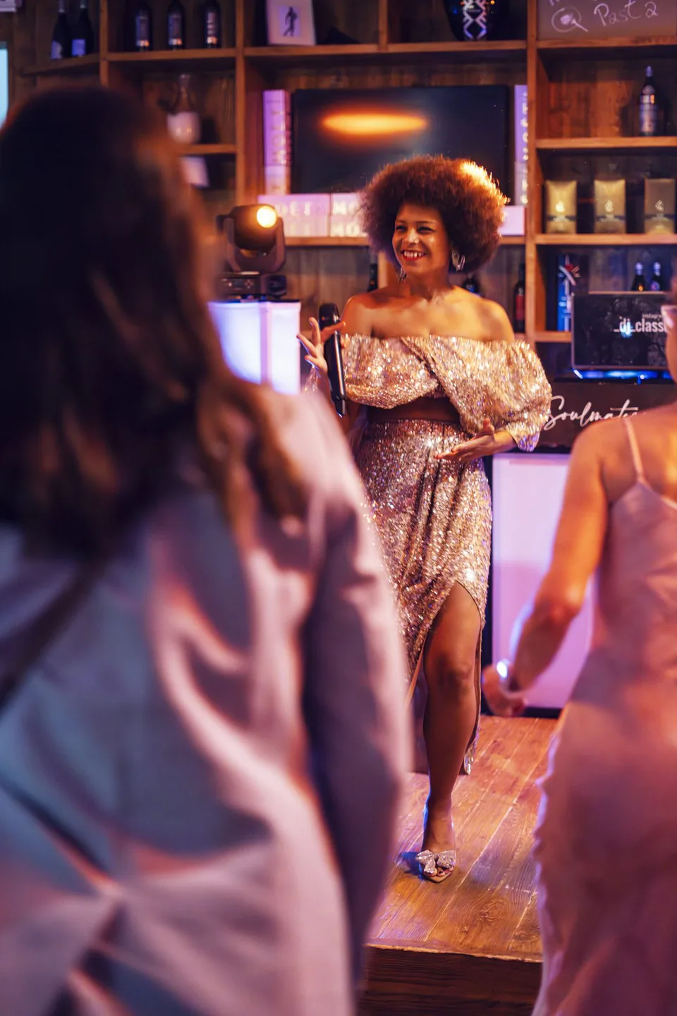 Frau im glitzernden Abendkleid hält ein Mikrofon vor Gästen in einer Bar mit warmem, violettem Licht.