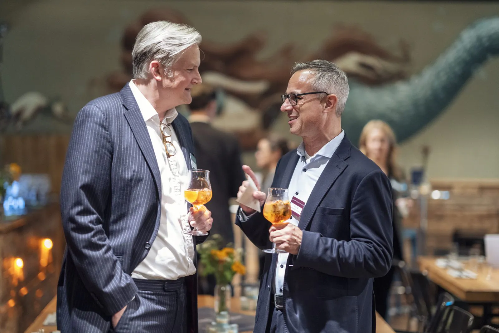 Zwei Männer in Anzügen unterhalten sich mit orangefarbenen Drinks in einem warm beleuchteten Veranstaltungsraum.