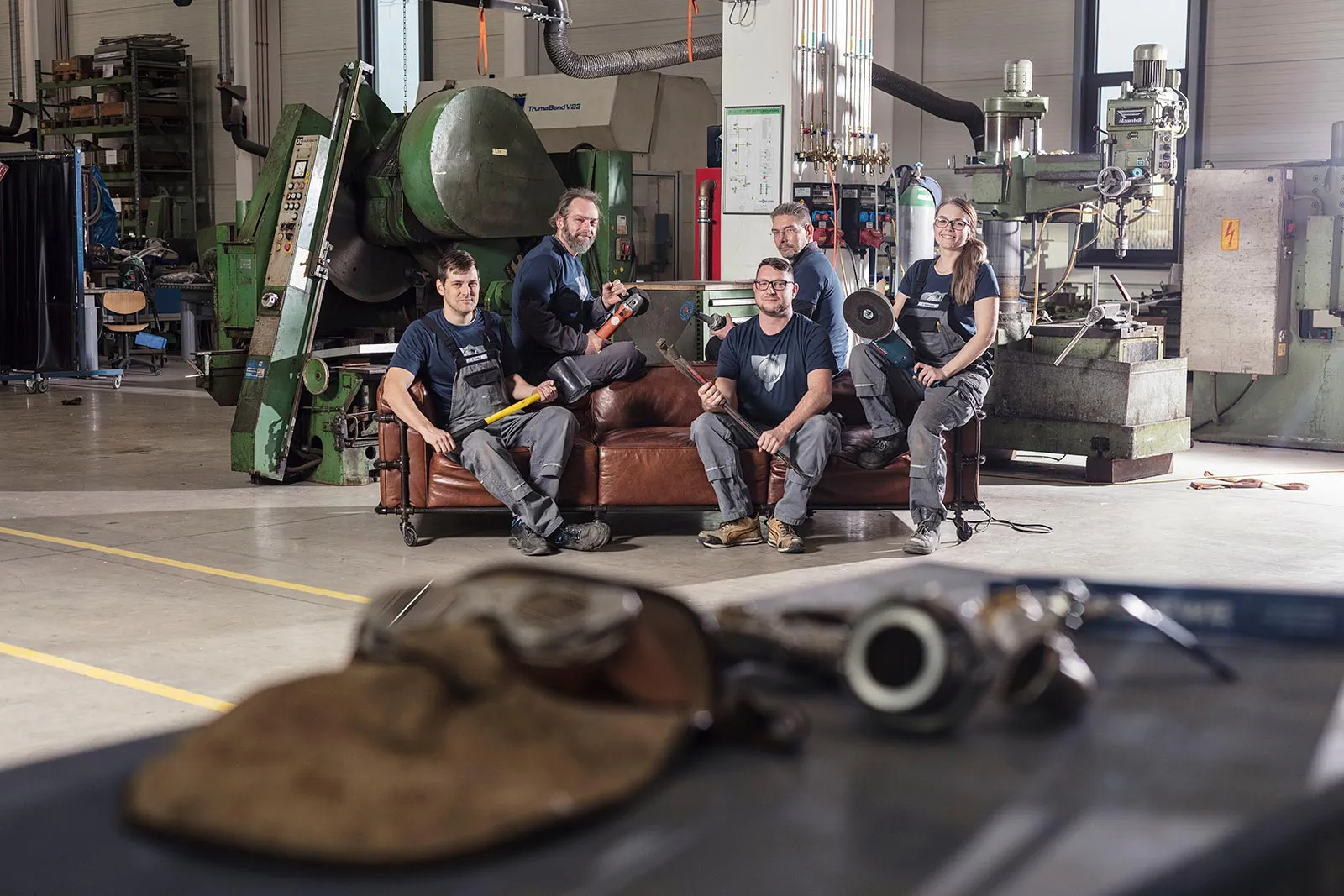 Fünf Personen mit Werkzeugen auf einem braunen Ledersofa in einer hellen Werkhalle mit großen Maschinen.