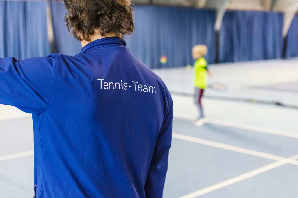 Person in blauer Tennis-Team-Jacke von hinten, dahinter ein Kind mit Tennisschläger in einer hellen Tennishalle.