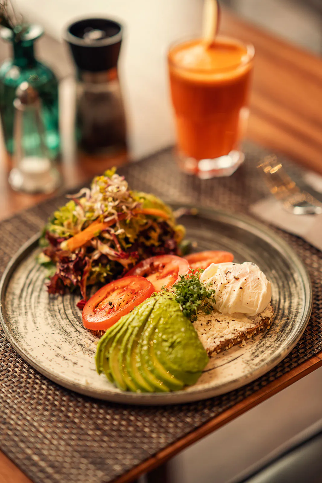 Teller mit Avocado, Tomatenscheiben, Blattsalat und einer weißen Beilage, dahinter ein Glas Saft im warmen Licht.