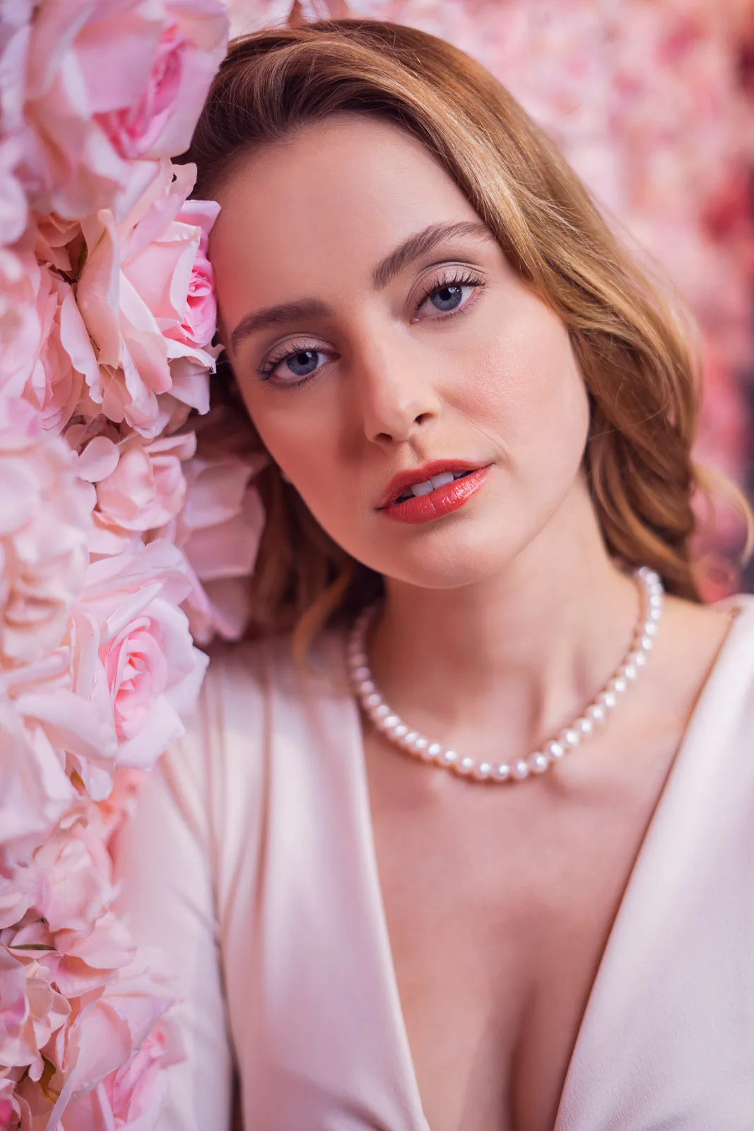 Frau mit Perlenkette und hellem Oberteil blickt im weichen Licht zwischen rosa Blüten in die Kamera.