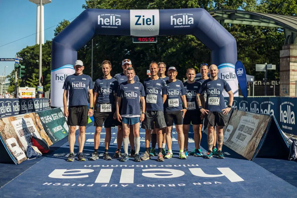 Läufergruppe in dunkelblauen Shirts posiert bei Sonnenschein unter dem hella-Zielbogen auf abgesperrter Straße.