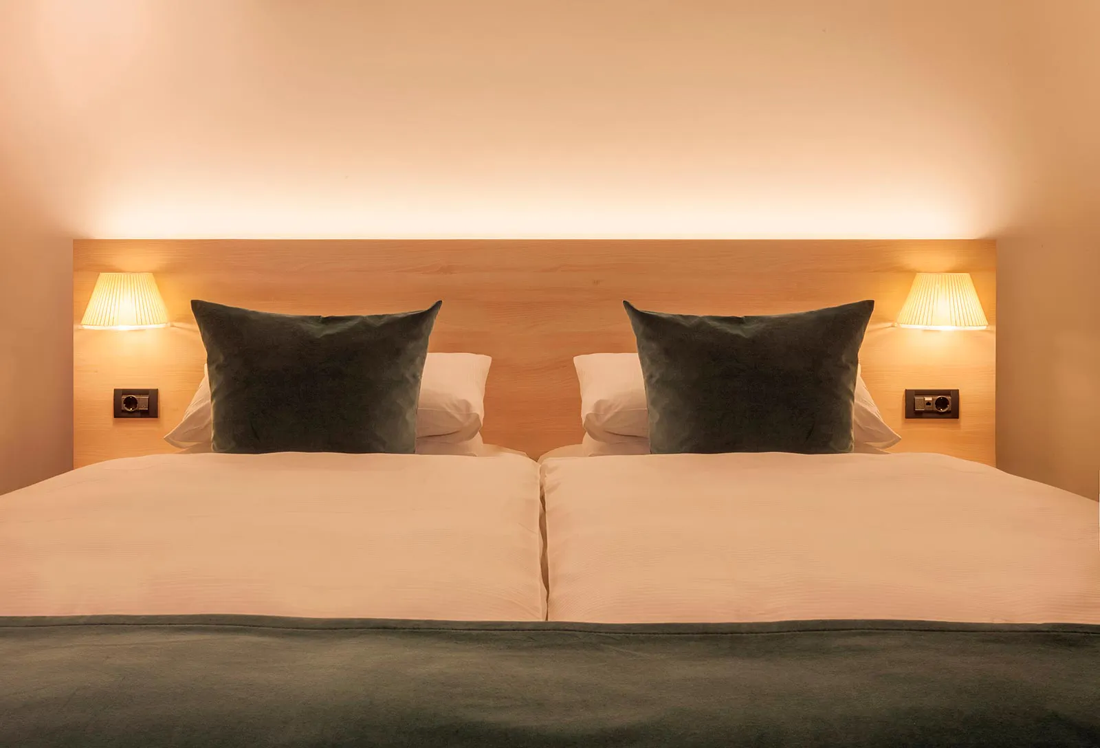 Doppelbett im Hotelzimmer mit weißer Bettwäsche, dunkelgrünen Kissen und warm beleuchtetem Holzkopfteil.