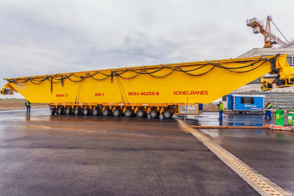 Gelber Konecranes-Kranträger auf Schwerlastfahrwerk, Arbeiter auf nassem Industriegelände unter grauem Himmel.