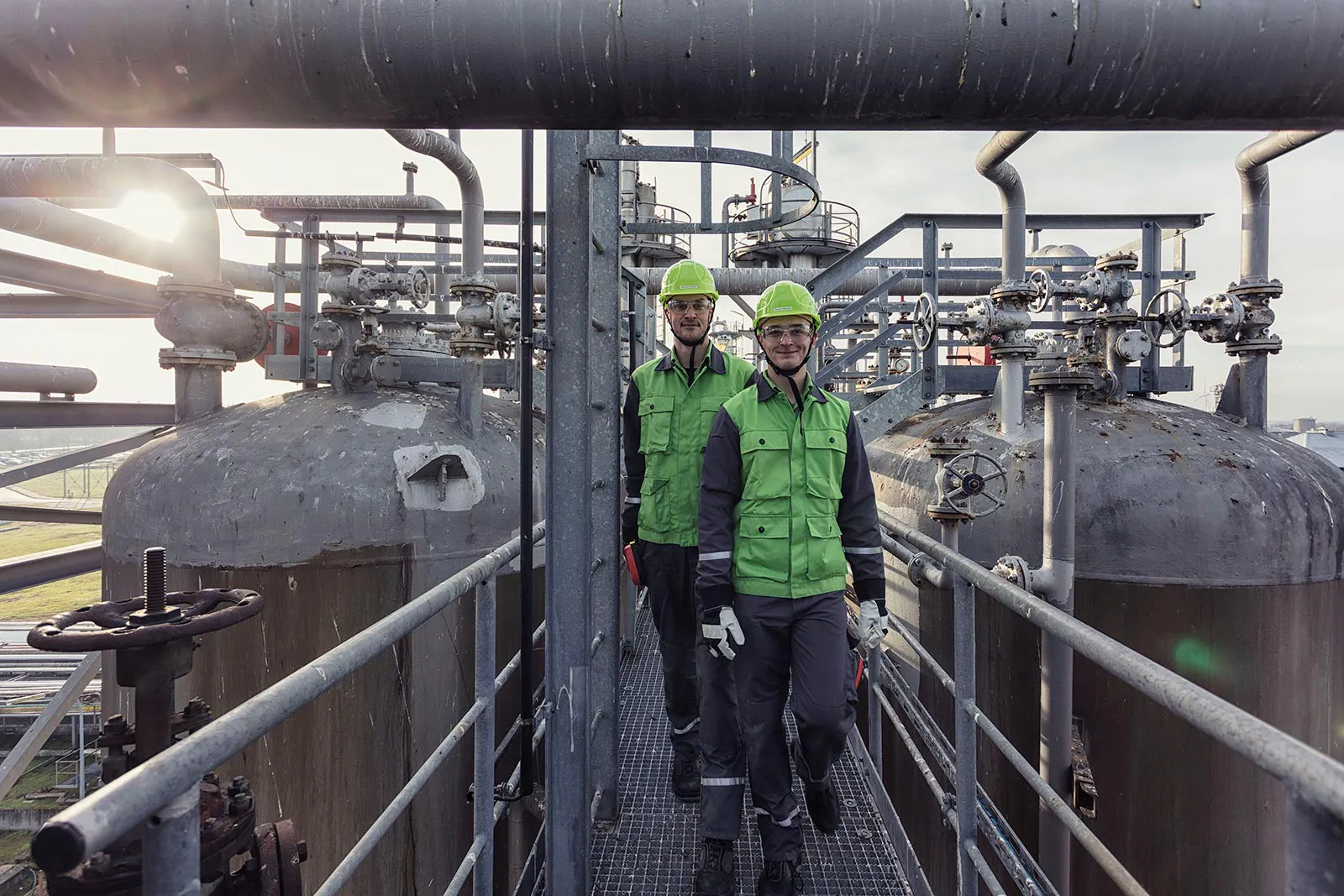 Zwei Personen mit grünen Helmen auf einem Metallsteg zwischen Rohren und Tanks der Raffinerie Heide im Gegenlicht.