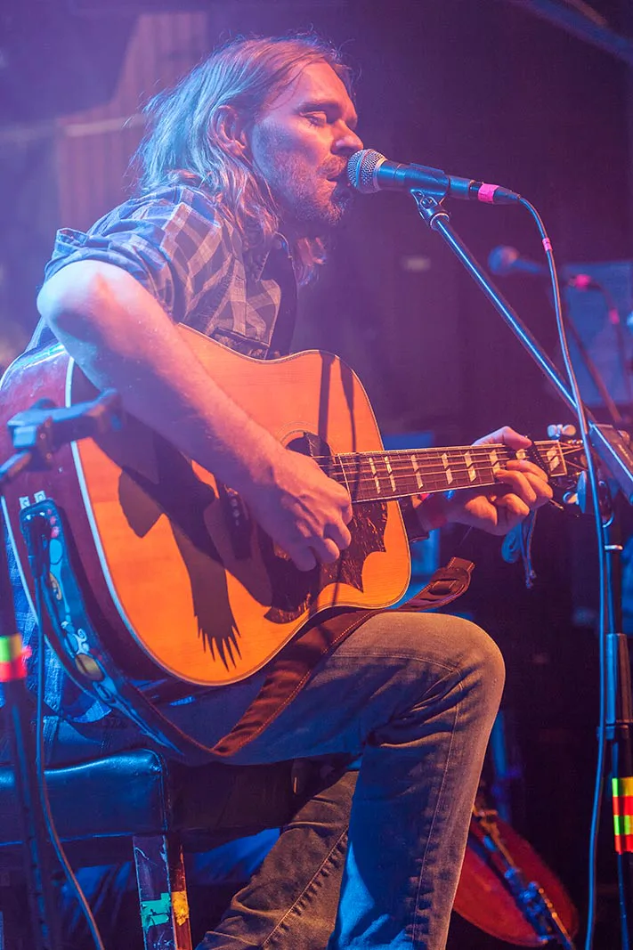 Sitzender Musiker mit Akustikgitarre singt ins Mikrofon auf einer blau und violett beleuchteten Bühne.