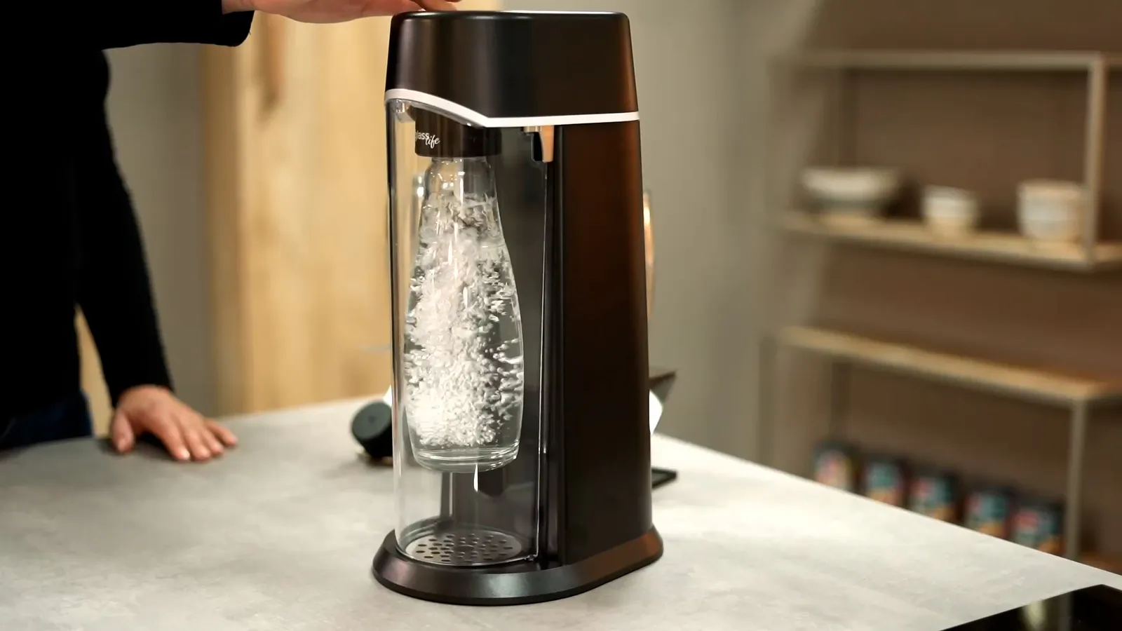 Person bedient einen schwarzen Wassersprudler mit sprudelnder Glasflasche auf einer Küchenarbeitsplatte bei warmem Licht.