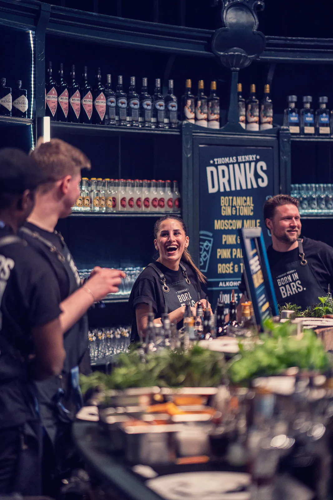 Lachendes Barteam hinter einer Theke mit Kräutern und Flaschen vor einem Thomas-Henry-Schild in dunkler Bar.