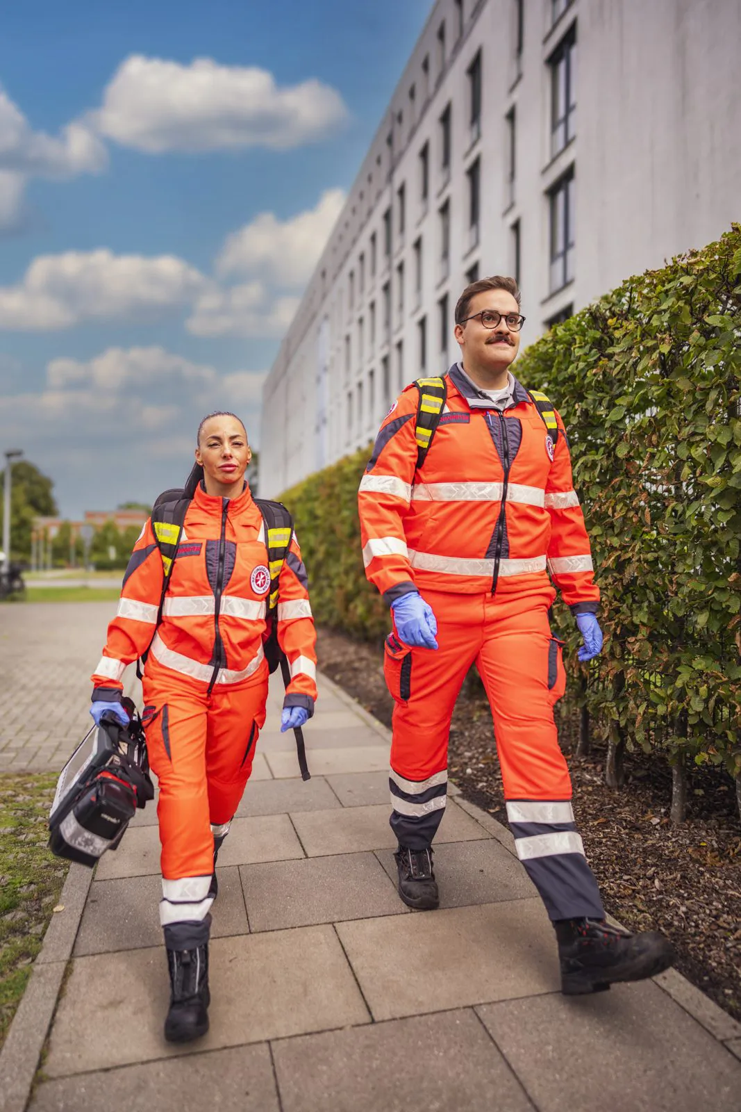 Zwei Johanniter-Rettungskräfte in orangefarbener Einsatzkleidung gehen bei Sonnenschein mit Ausrüstung vor einem Gebäude.