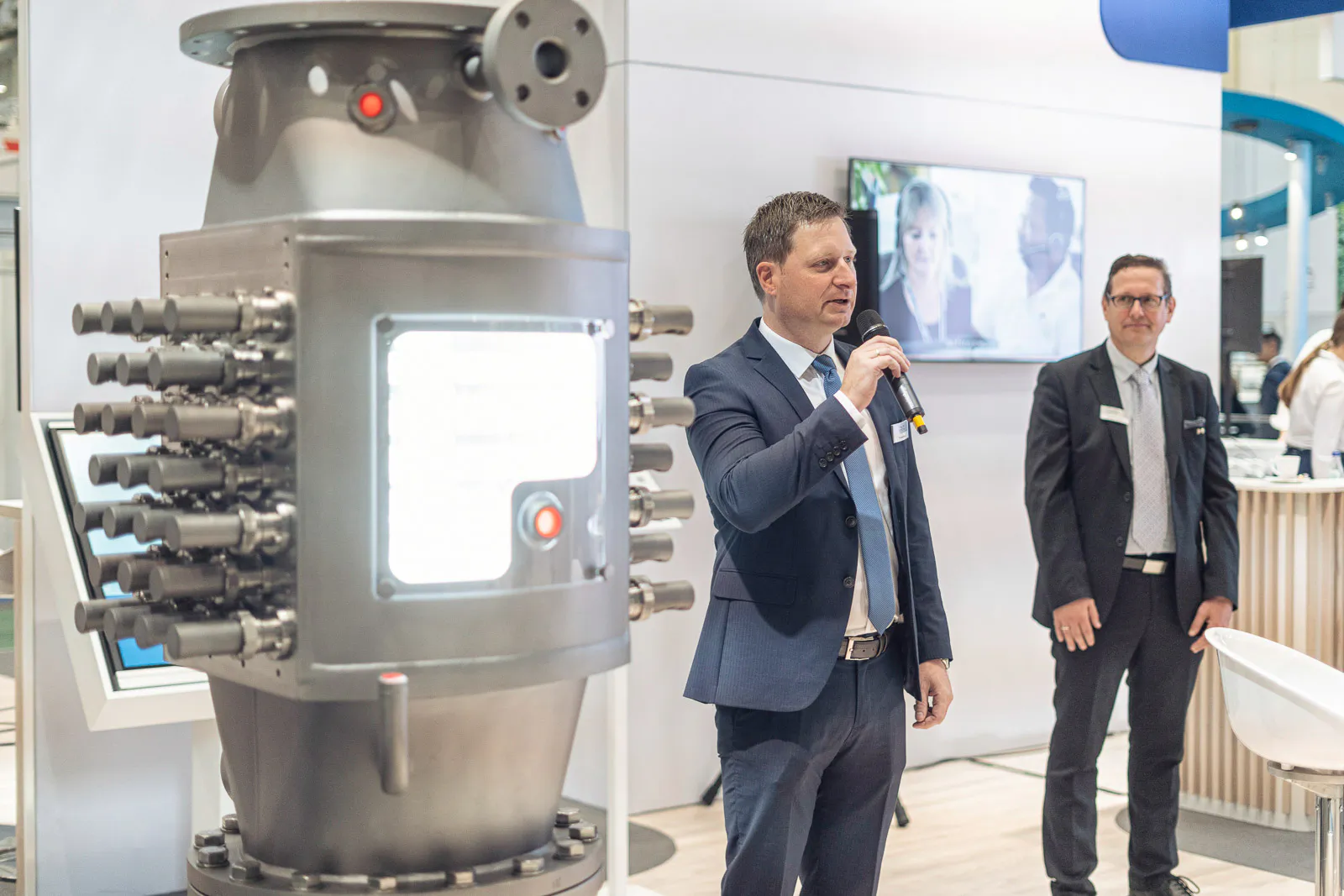 Mann mit Mikrofon neben einer großen Metallanlage am hellen Alfa-Laval-Messestand, rechts ein weiterer Mann im Anzug.
