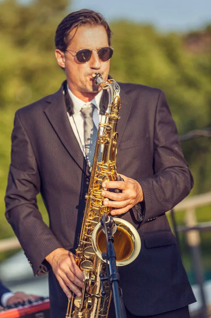 Saxofonist im dunklen Anzug mit Sonnenbrille spielt im warmen Sonnenlicht im Freien vor einem Mikrofon.