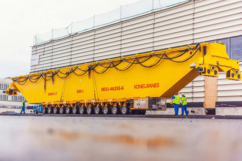Gelber Konecranes-Kranträger auf Schwerlastfahrwerk vor einer Industriehalle, daneben Arbeiter in Warnkleidung.