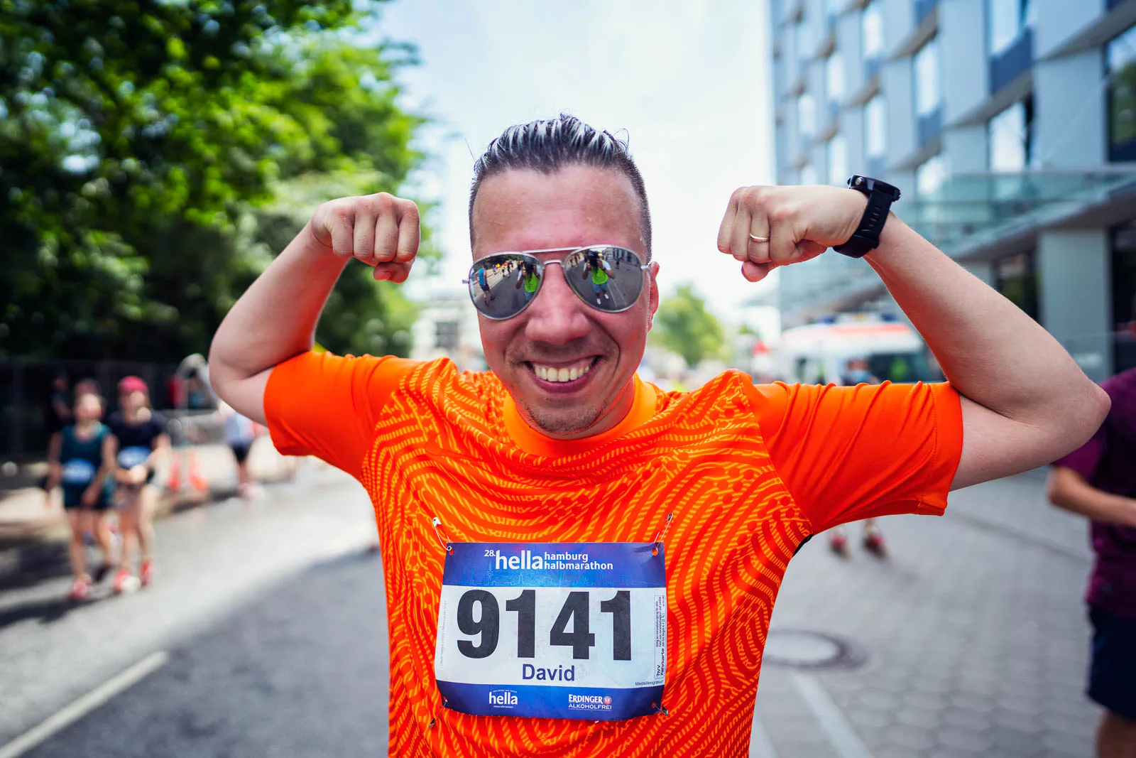 Läufer im orangefarbenen Shirt mit Startnummer 9141 und Sonnenbrille zeigt lächelnd seine Muskeln auf sonniger Straße.