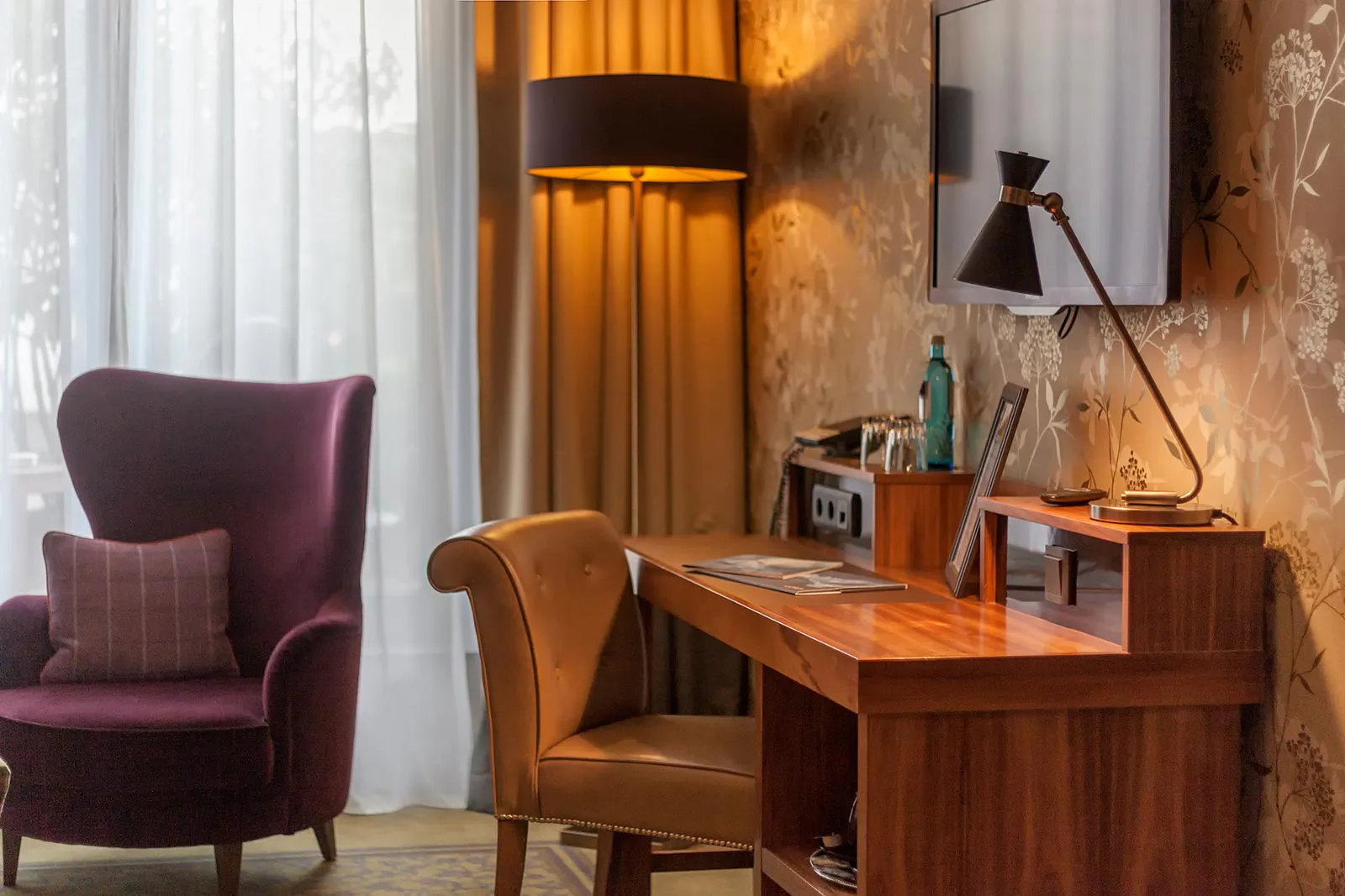 Hotelzimmer mit violettem Sessel, Holzschreibtisch, Lederstuhl und warmem Lampenlicht neben einem hellen Fenster.