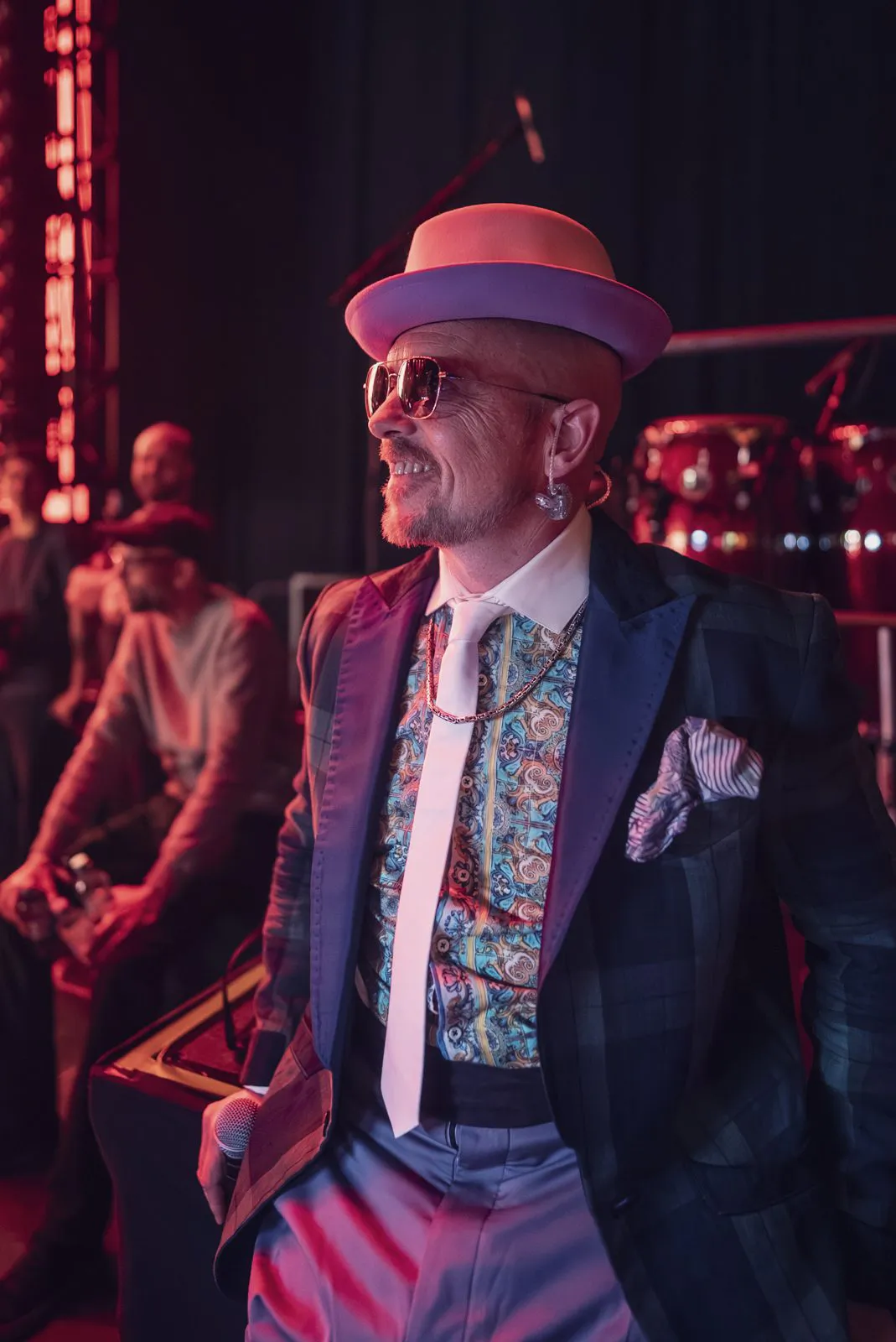 Jan Delay mit Hut, Sonnenbrille und Mikrofon im roten Bühnenlicht, dahinter Musiker und Trommeln.