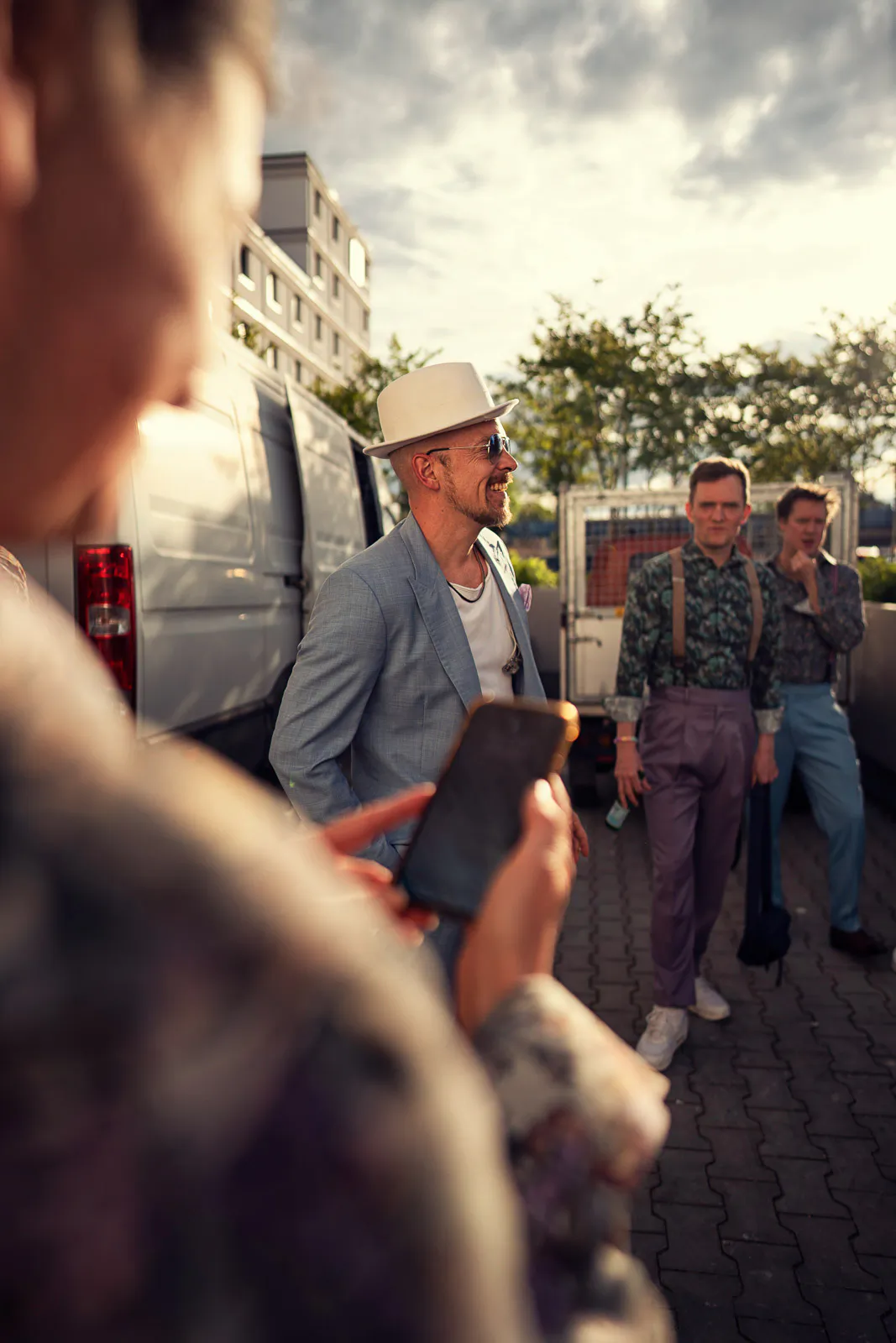 Jan Delay mit Hut, Sonnenbrille und grauem Sakko neben einem Transporter, umringt von Menschen im warmen Abendlicht.