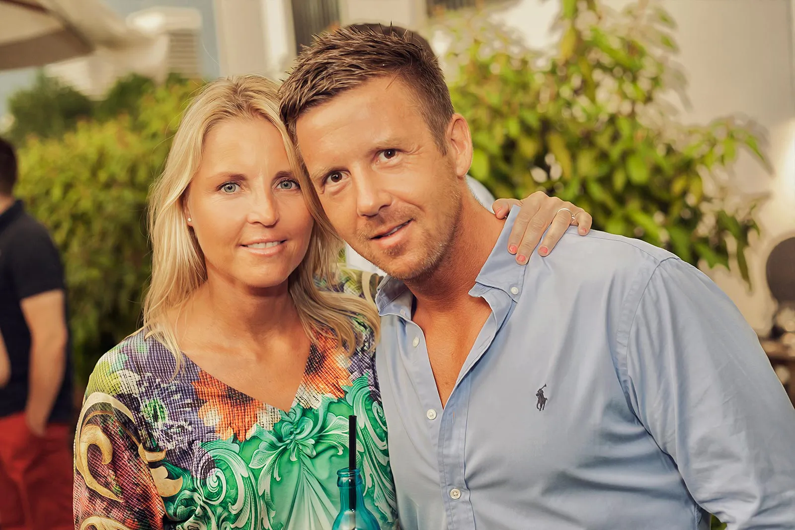 Frau im bunten Kleid und Mann im hellblauen Hemd posieren eng beieinander auf einer sonnigen Terrasse.