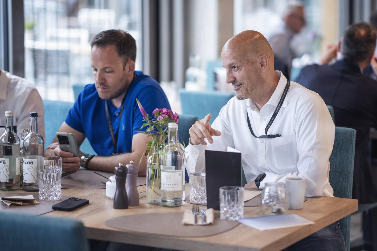 Zwei Männer sitzen im hellen Restaurant am gedeckten Tisch; einer schaut aufs Handy, der andere gestikuliert.