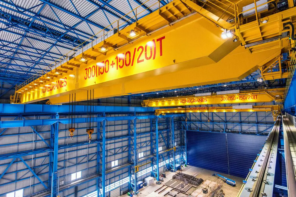 Gelbe Konecranes-Brückenkrane unter beleuchtetem Hallendach mit blauen Stahlträgern in einer Industriehalle.