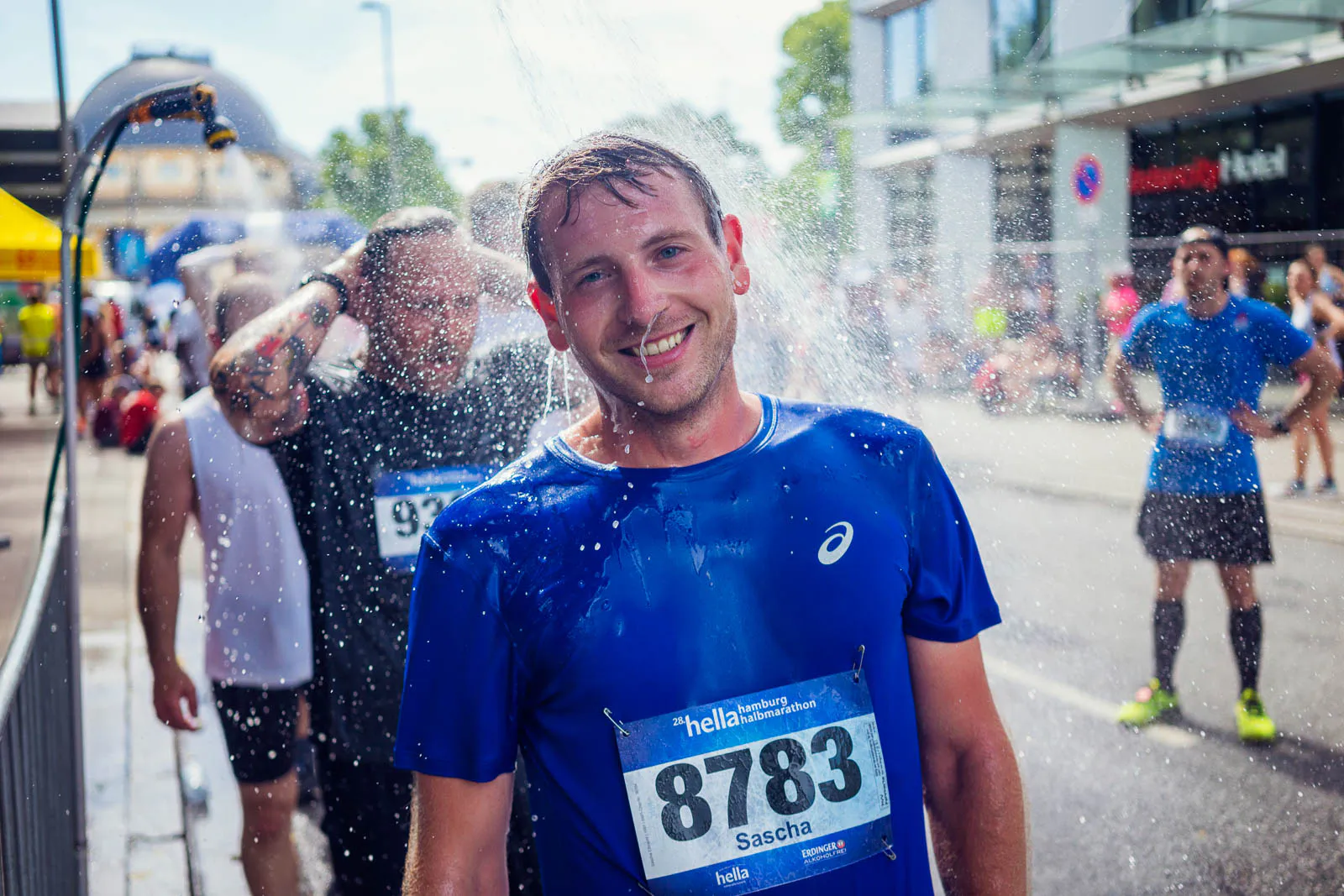 Lächelnder Läufer im blauen Shirt mit Startnummer unter einer Wasserdusche auf sonniger Straße beim Hella Halbmarathon.