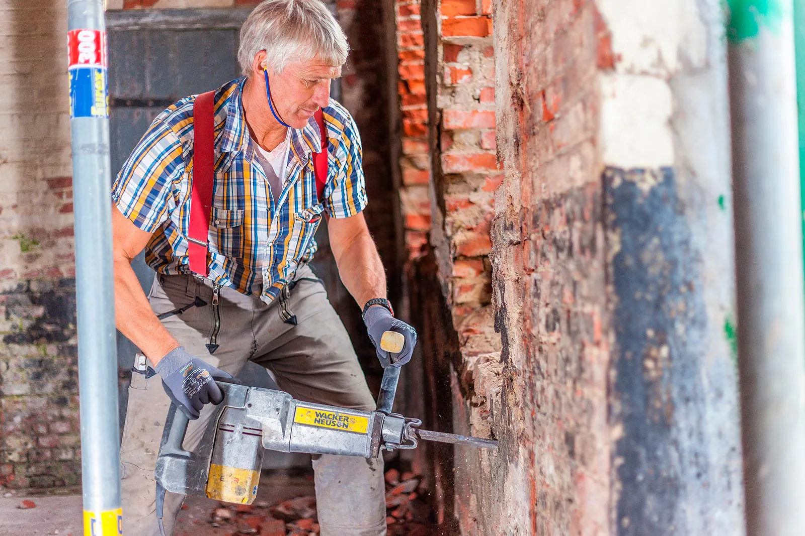 Handwerker mit Handschuhen und Abbruchhammer bearbeitet eine freigelegte Ziegelwand auf einer hellen Baustelle.