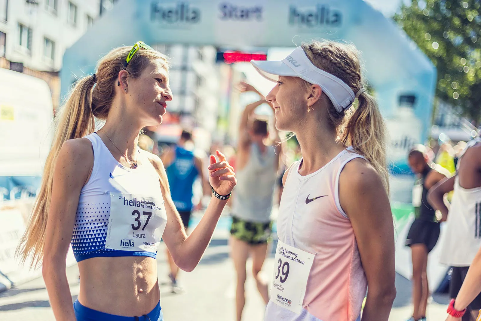 Zwei Läuferinnen mit Startnummern unterhalten sich im Sonnenlicht vor dem Startbogen des Hella Halbmarathons.