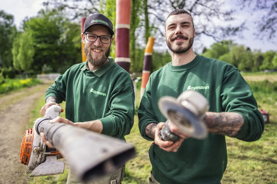 Zwei lächelnde Männer in grüner Boymann-Arbeitskleidung halten Gartengeräte im sonnigen Grün.