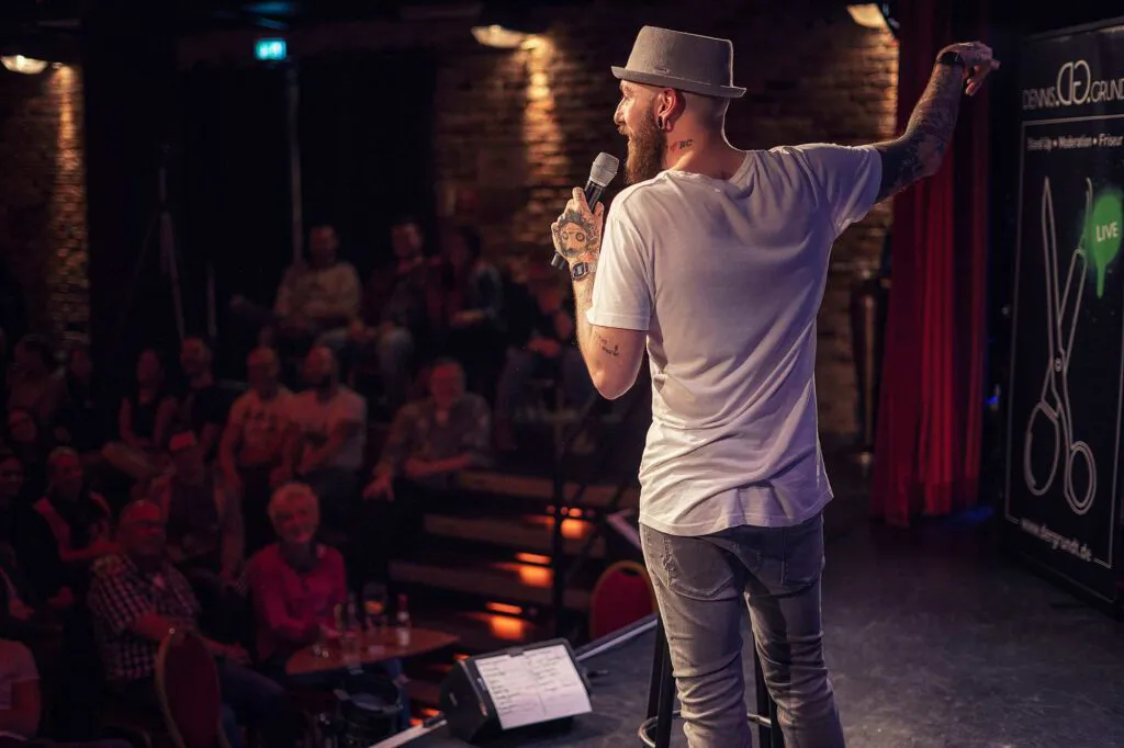 Dennis Grundt mit Hut und Mikrofon auf einer warm beleuchteten Clubbühne vor Publikum, einen Arm erhoben.