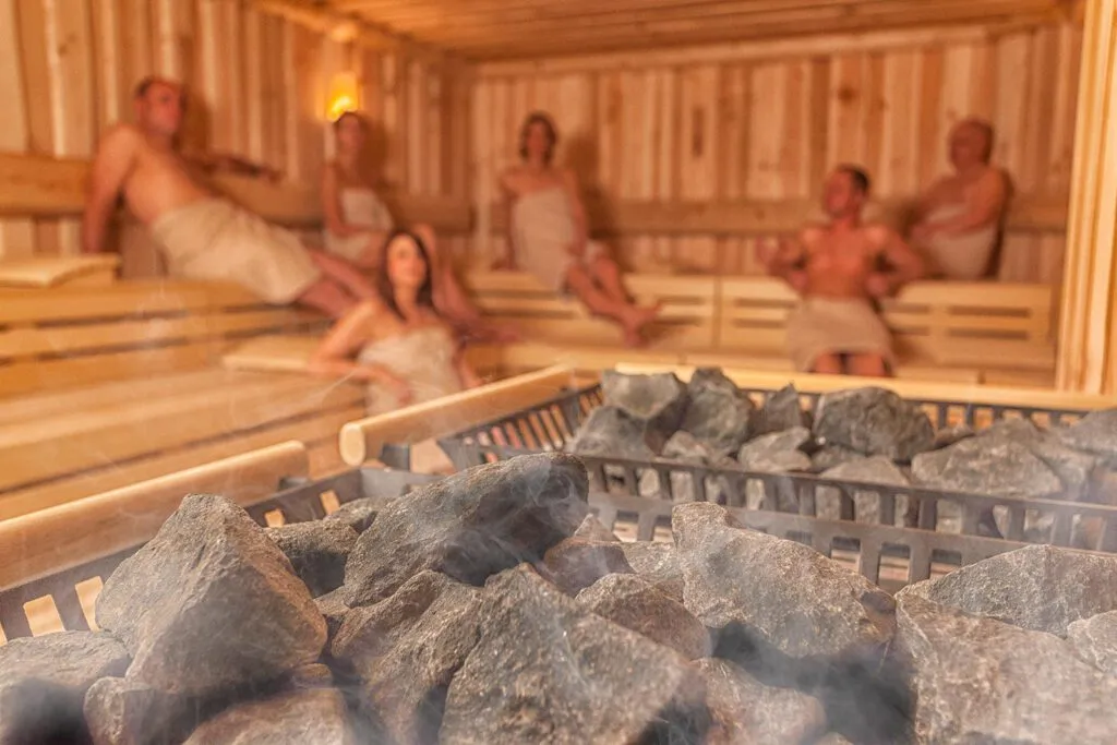 Sechs Gäste mit Handtüchern in einer warm beleuchteten Holzsauna, Saunasteine im Vordergrund.