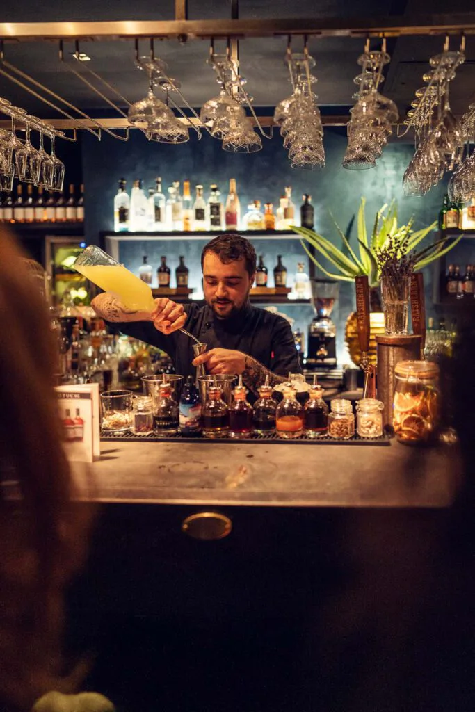 Barkeeper gießt gelbe Flüssigkeit in einen Messbecher hinter einer warm beleuchteten Bar mit Flaschen und Hängegläsern.