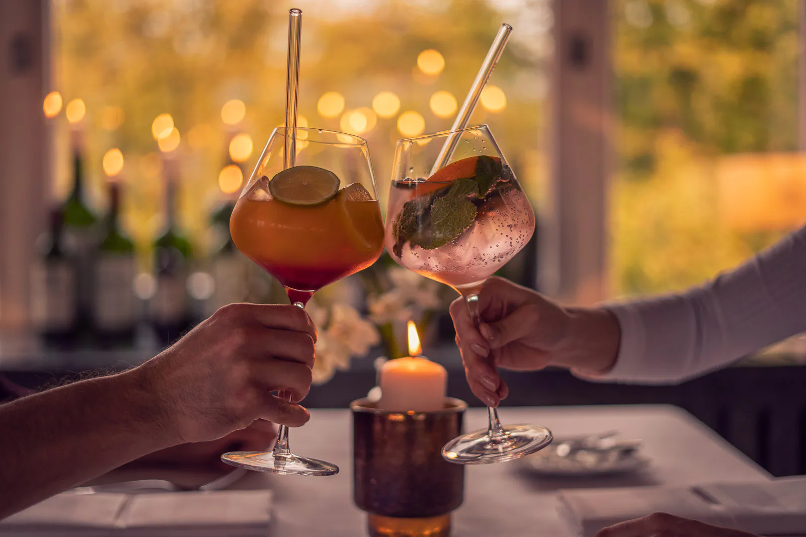 Zwei Personen stoßen mit bunten Cocktails in Stielgläsern an, im warmen Kerzenlicht an einem gedeckten Tisch.