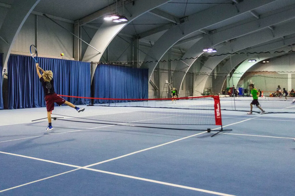 Kind beim Aufschlag am Wilson-Tennisnetz in einer beleuchteten Halle, gegenüber ein Kind im grünen Shirt.
