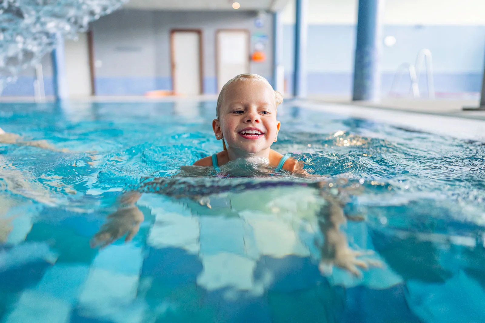 Lächelndes Kind schwimmt im türkisblauen Wasser eines hellen Hallenbads, die Arme unter Wasser ausgestreckt.