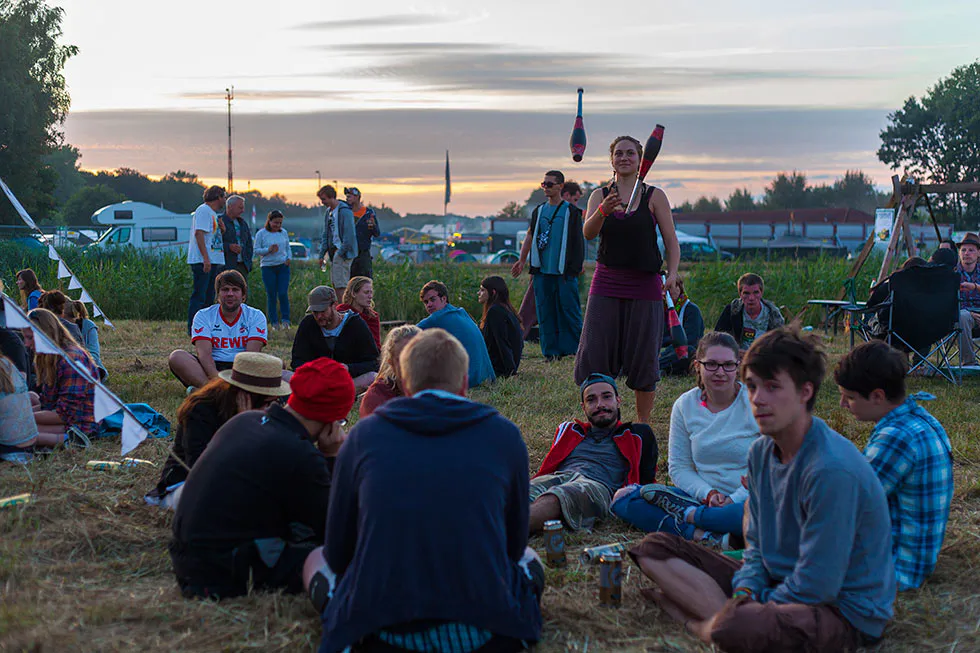 Eine Person jongliert mit Keulen, während Festivalgäste bei Sonnenuntergang auf einer Wiese sitzen.