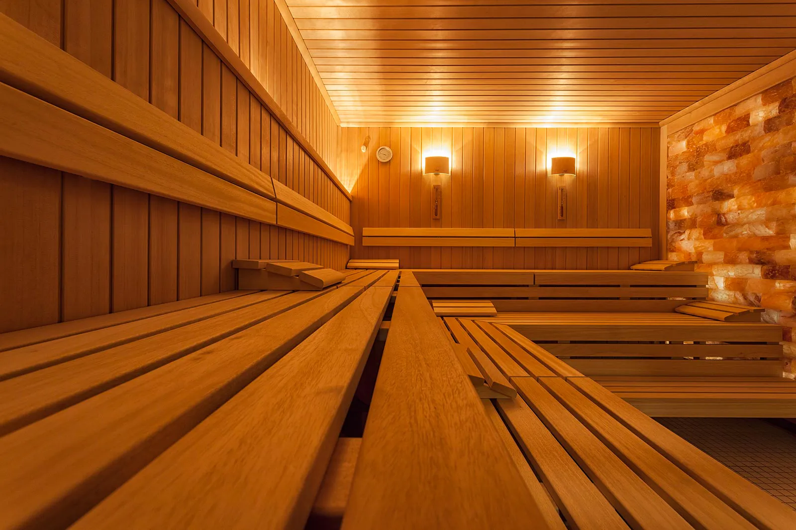 Leere Sauna mit mehrstufigen Holzbänken, warmen Wandleuchten und einer Wand aus orangefarbenen Salzsteinen.