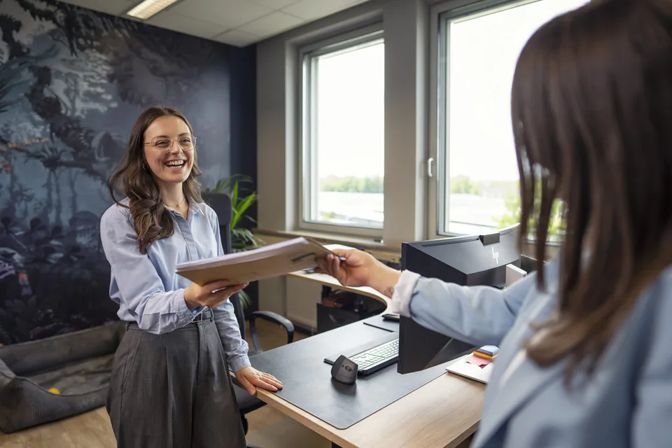 Zwei Frauen übergeben eine Mappe über einen Büroschreibtisch; Tageslicht fällt durch große Fenster.