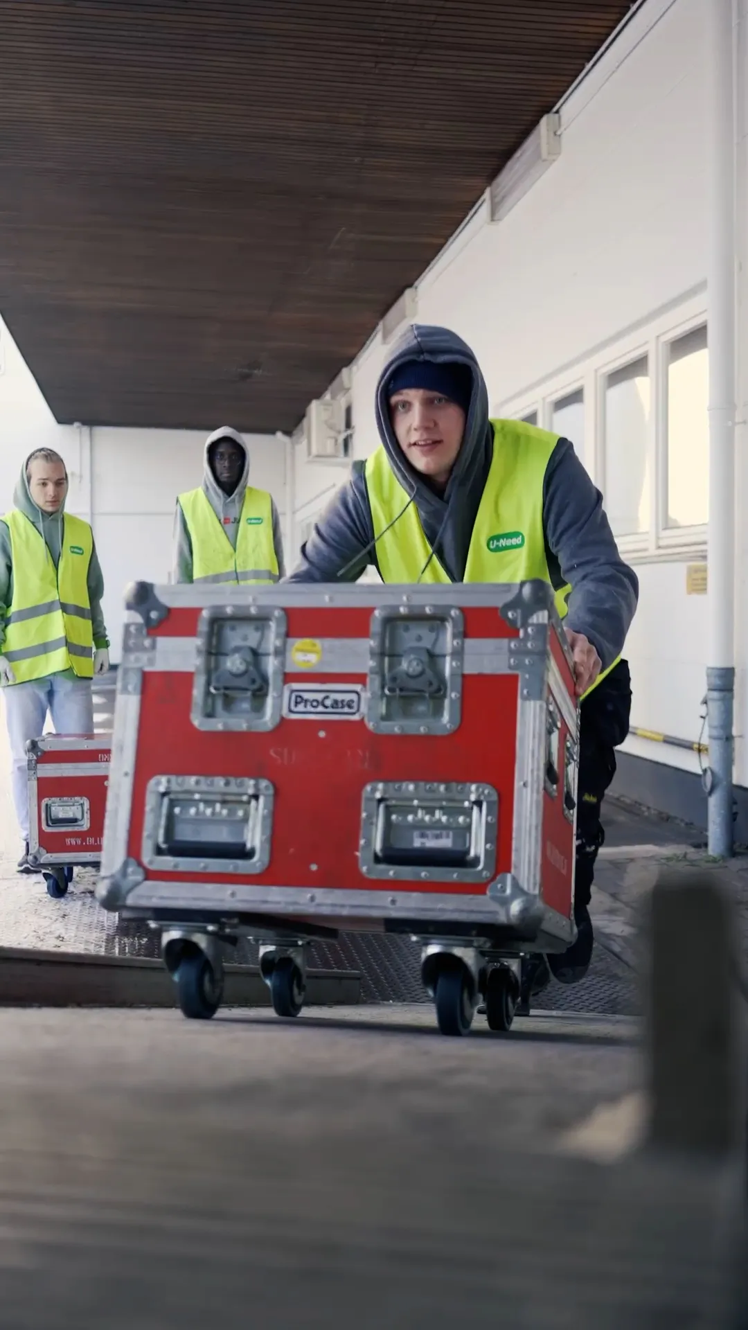 Drei Personen in gelben Warnwesten schieben rote Transportkisten auf Rollen durch einen überdachten Gang.