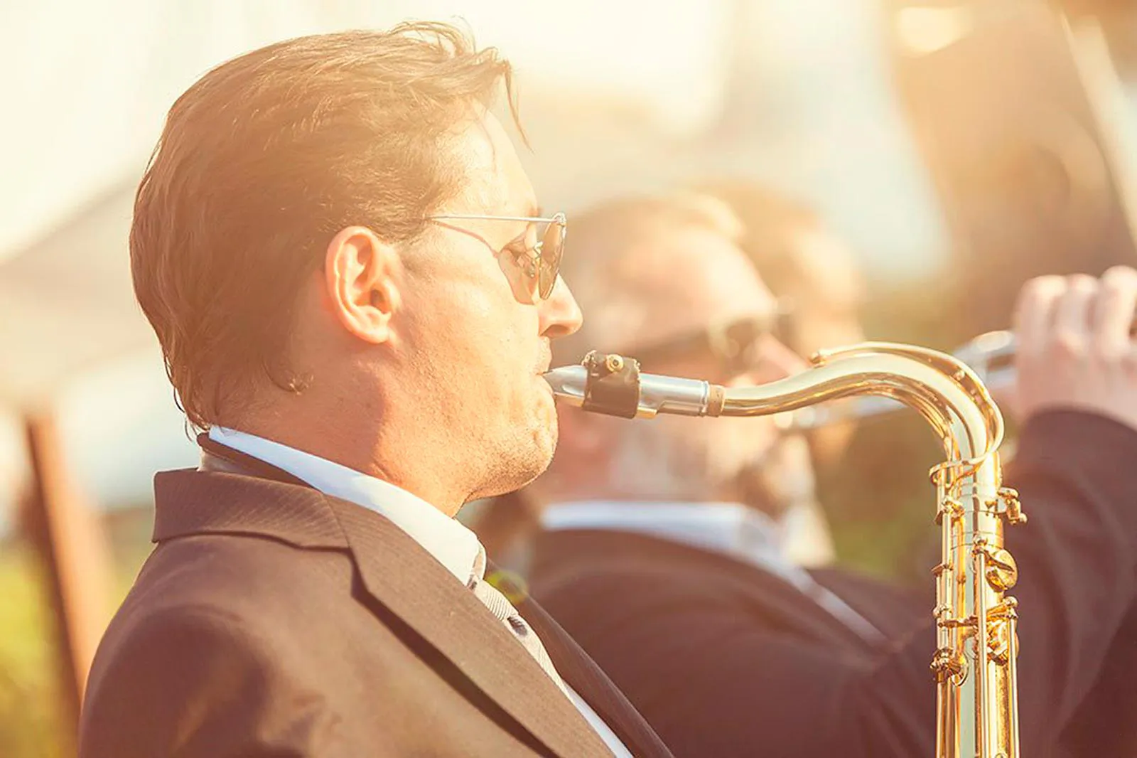 Musiker im Anzug mit Sonnenbrille spielt Saxofon im warmen Sonnenlicht, dahinter ein Trompeter.