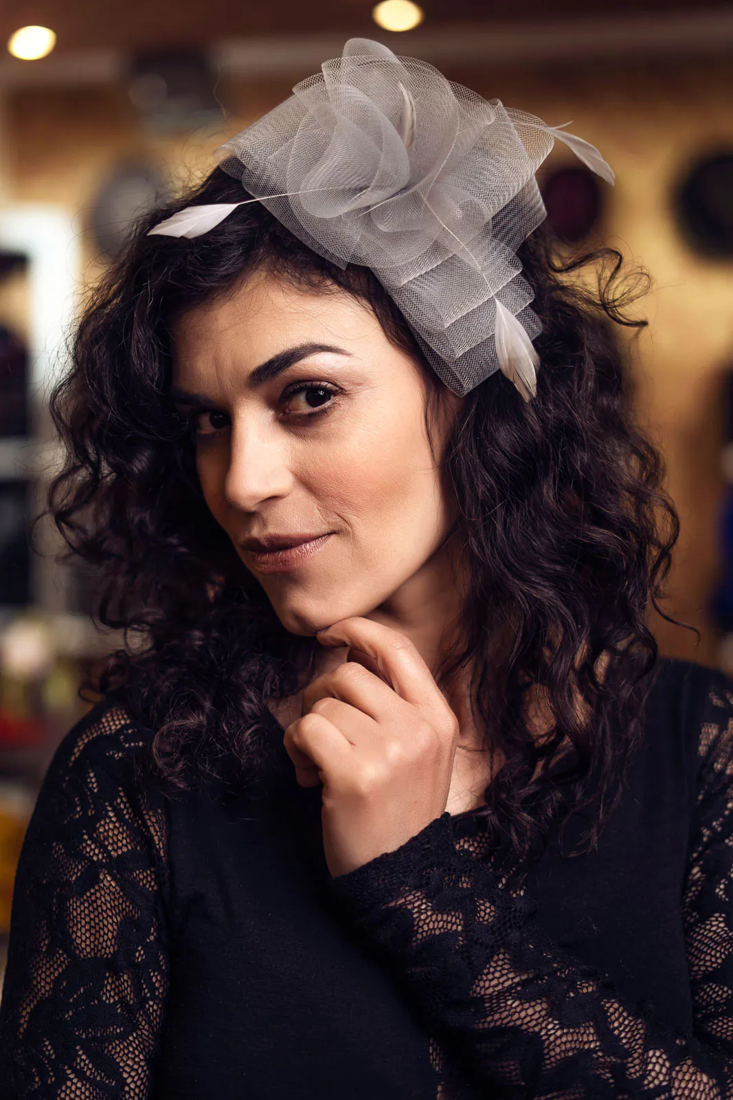 Frau mit dunklen Locken und grauem Fascinator hält die Hand ans Kinn, in warm beleuchtetem Laden.