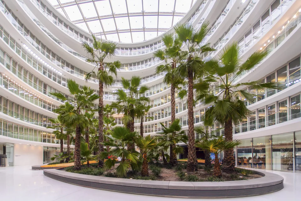 Lichtdurchflutetes Atrium mit hohen Palmen, geschwungenen Fensterreihen, Glasdach und umlaufender Sitzbank.
