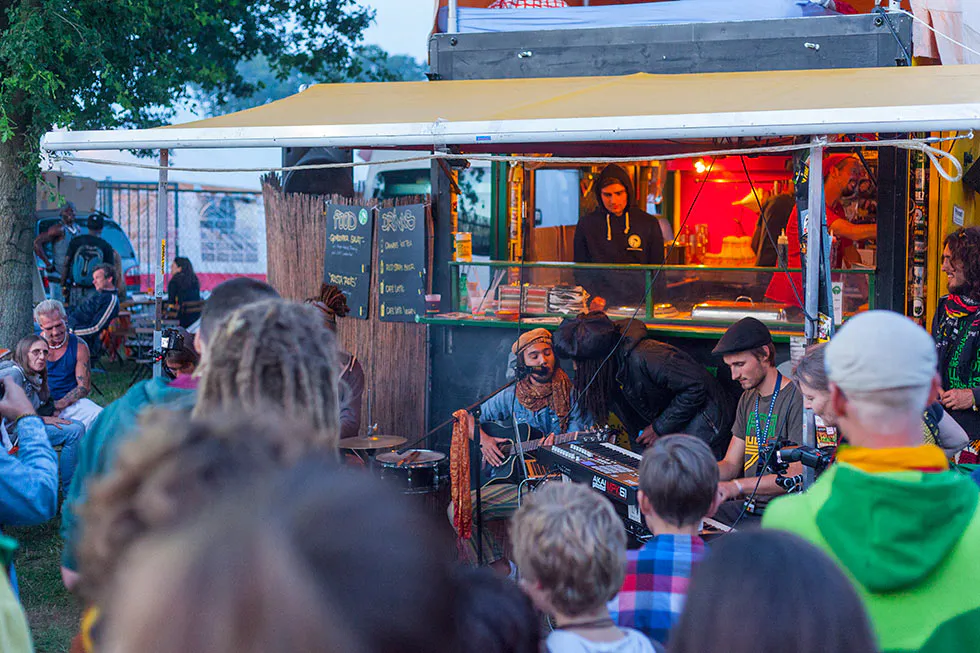 Musiker mit Gitarre und Mischpult vor einem rot beleuchteten Imbisswagen, umringt von Festivalbesuchern.