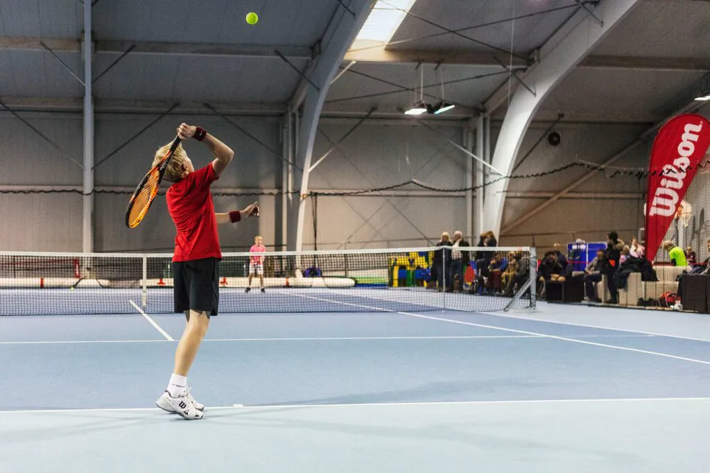 Tennisspieler im roten Shirt beim Aufschlag auf blauem Hallenplatz, Zuschauer und rote Wilson-Fahne im Hintergrund.