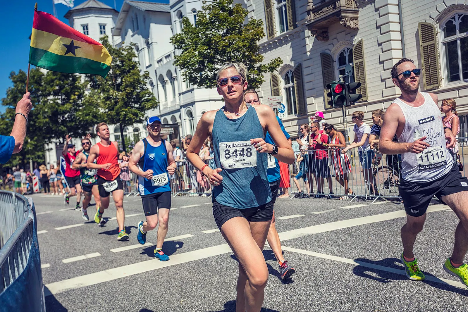 Läufer beim Hella Halbmarathon auf sonniger Straße, Zuschauer hinter Absperrungen und eine Ghana-Flagge am Rand.