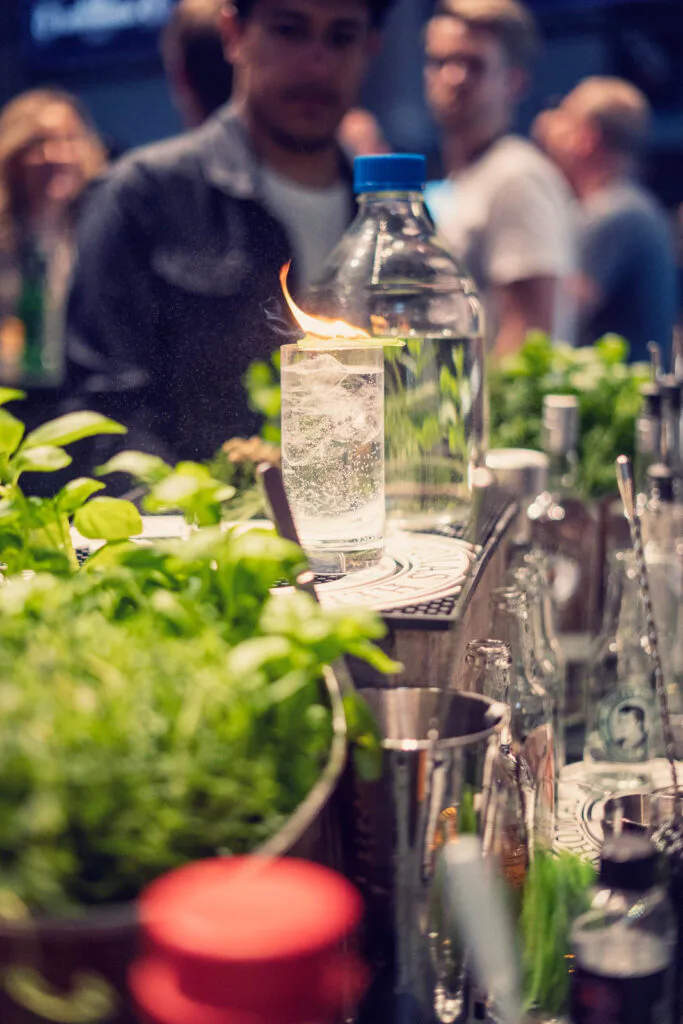 Flammender Drink im hohen Glas auf einer Bar mit frischen Kräutern, dahinter Gäste im gedämpften Eventlicht.