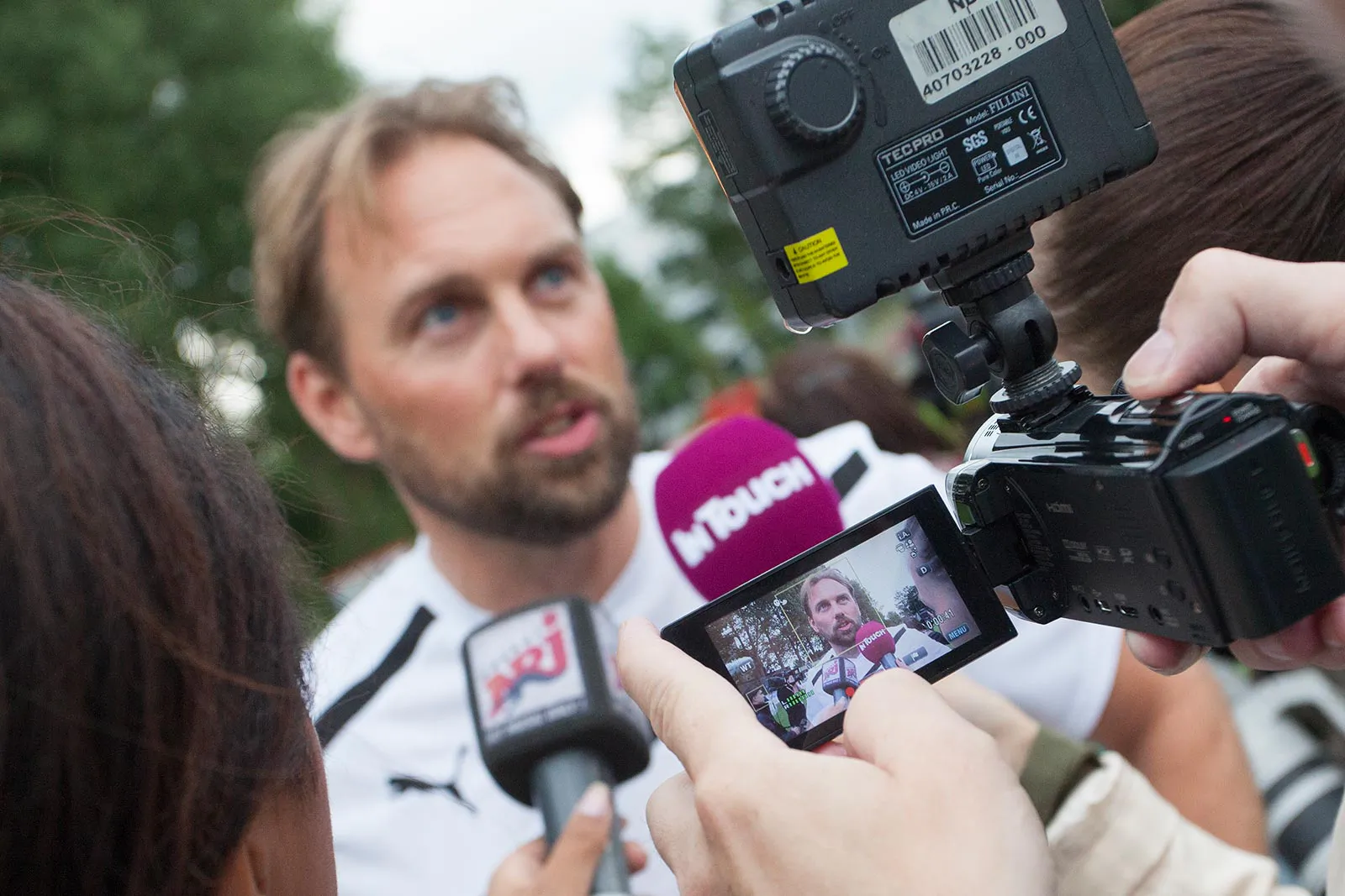 Mann im weißen Sportshirt gibt draußen ein Interview vor mehreren Mikrofonen und einer Videokamera mit LED-Leuchte.